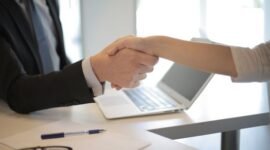 Dua pebisnis berjabat tangan di atas meja dengan dokumen dan grafik, melambangkan momen krusial dalam proses merger dan akuisisi yang strategis. Jabat tangan ini menandai kesepakatan penting yang akan membentuk arah masa depan bisnis kedua belah pihak.