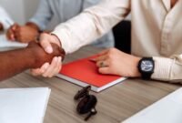 Dua pebisnis berjabat tangan di atas latar belakang gedung pencakar langit kota, melambangkan kesepakatan merger yang strategis untuk mencapai tujuan bisnis yang lebih besar. Penggabungan ini diharapkan membawa sinergi dan pertumbuhan signifikan bagi kedua belah pihak.