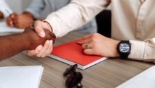 Dua pebisnis berjabat tangan di atas latar belakang gedung pencakar langit kota, melambangkan kesepakatan merger yang strategis untuk mencapai tujuan bisnis yang lebih besar. Penggabungan ini diharapkan membawa sinergi dan pertumbuhan signifikan bagi kedua belah pihak.